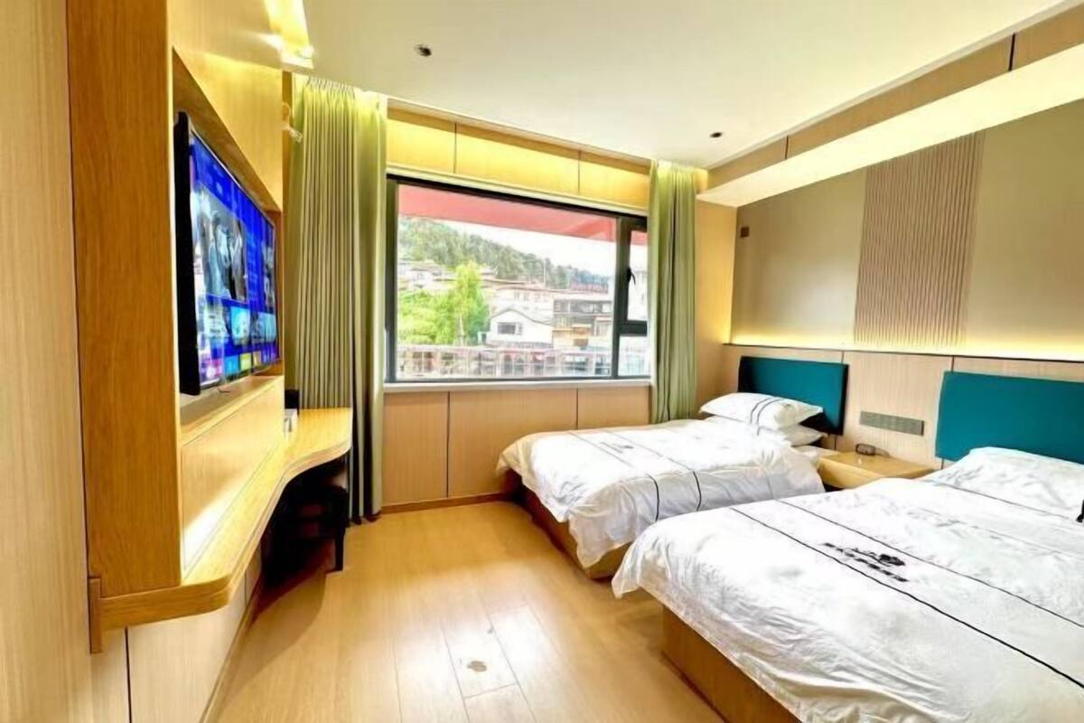 Una habitación de hotel con dos camas, televisión, ventana con cortinas y vista a edificios.