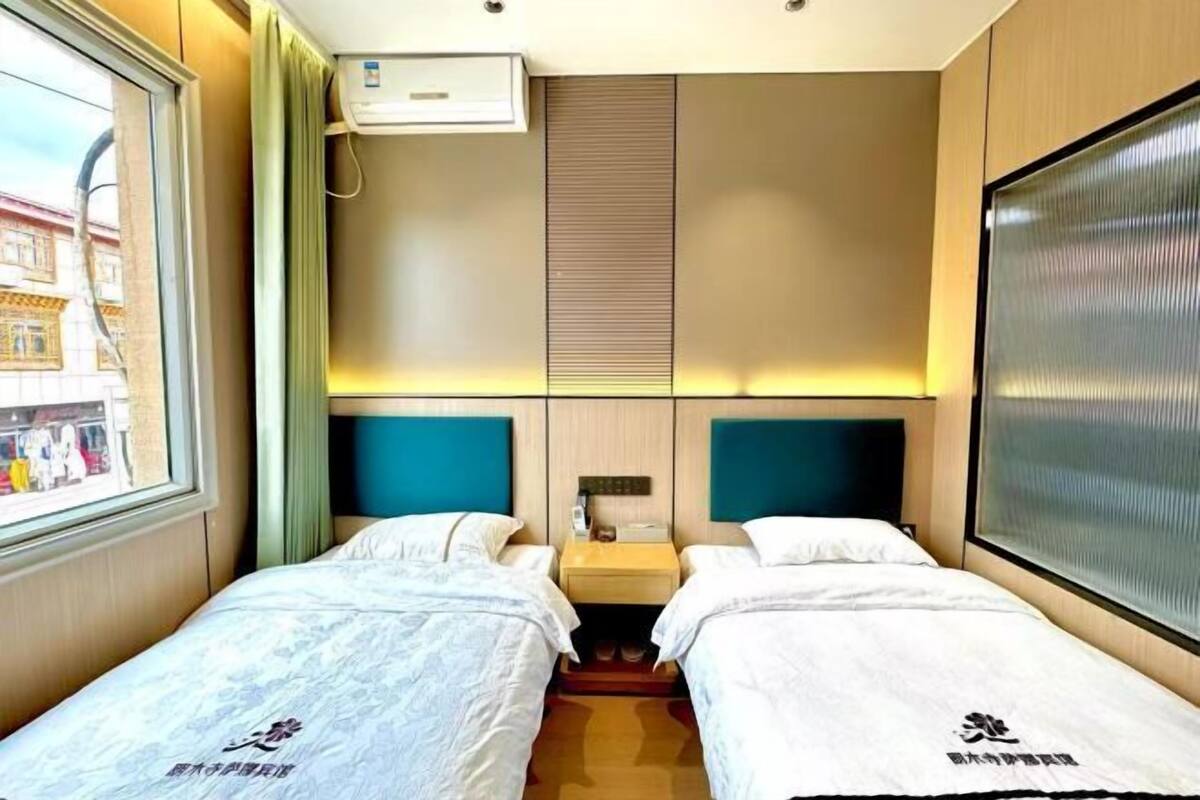 Habitación de hotel con dos camas, una ventana con vistas a edificios, un aire acondicionado de pared y una mesita de noche con lámpara.