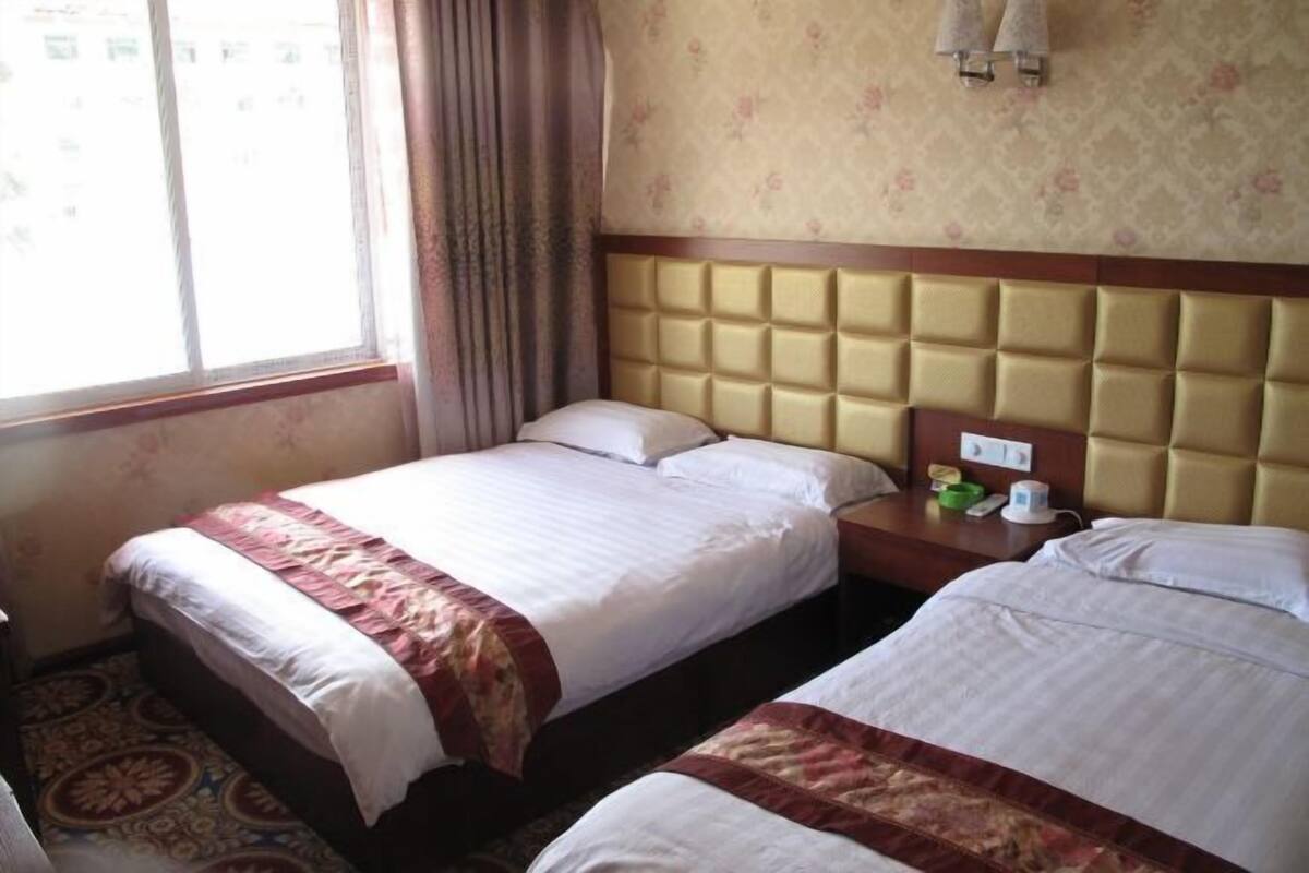 Habitación de hotel con dos camas, cabecero, mesita de noche, ventana con cortinas y alfombra estampada.
