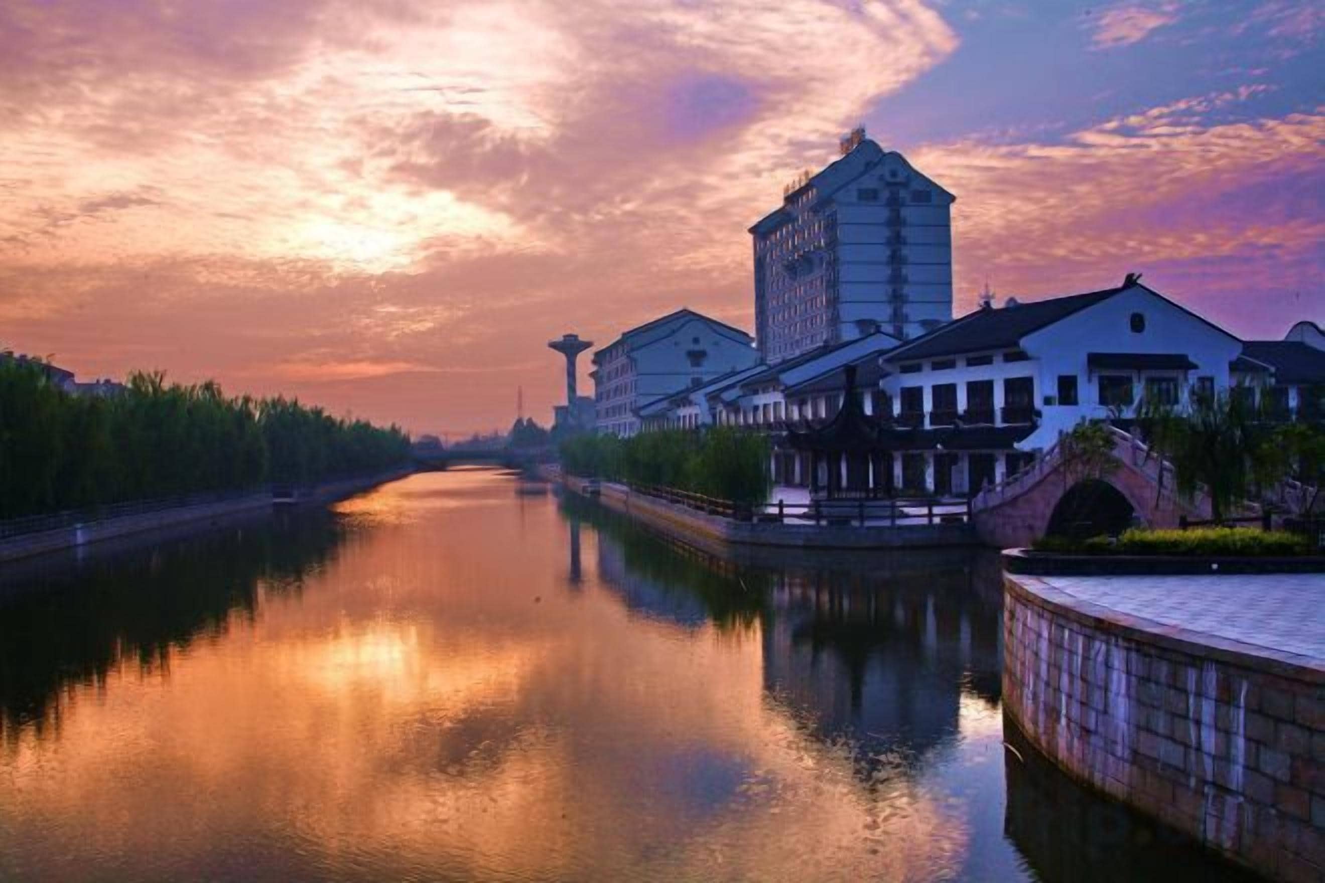 Een rustige rivier met rechts gebouwen, een brug en een zonsondergang.