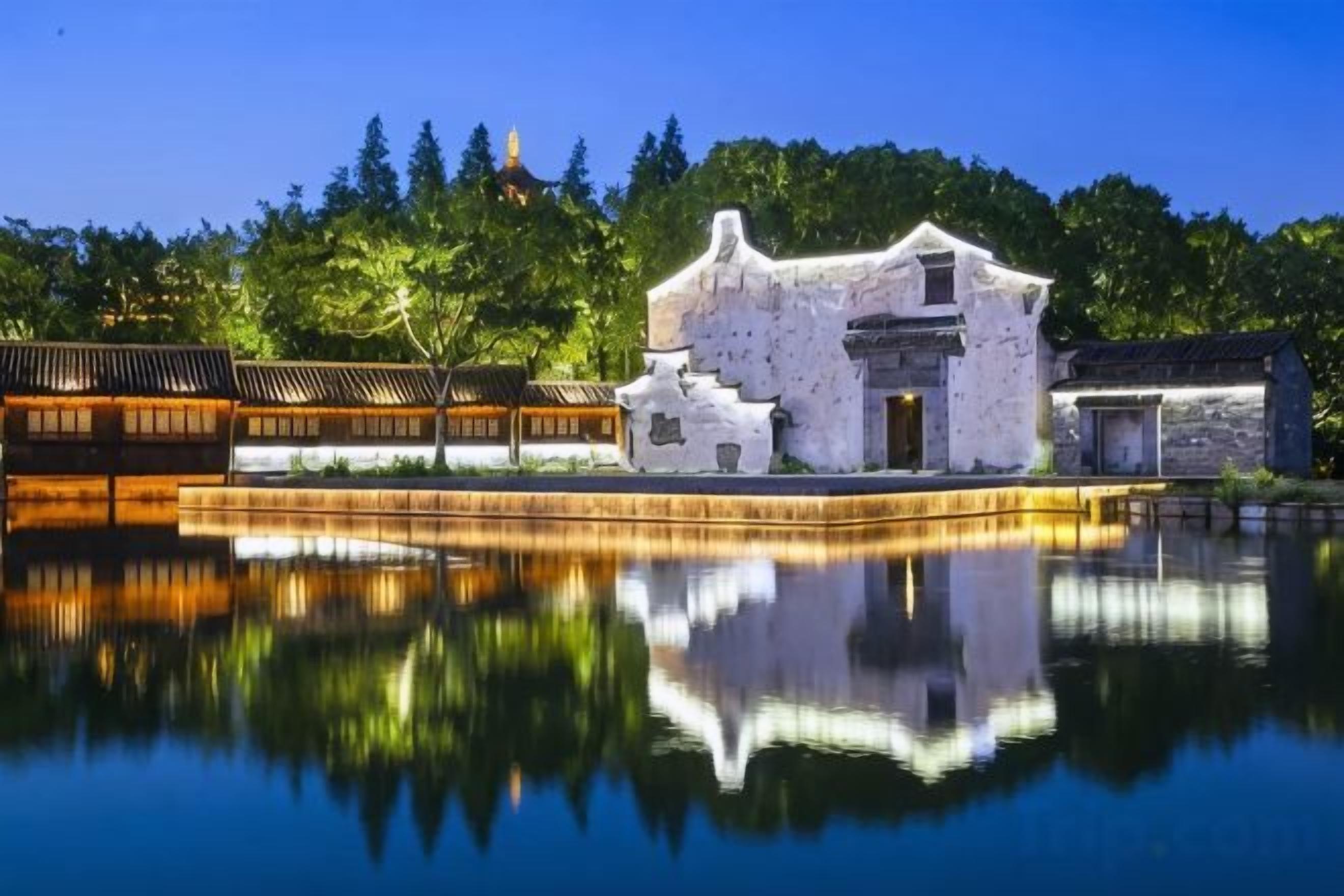 A traditional building with a reflection in a calm body of water, surrounded by trees and a clear sky.