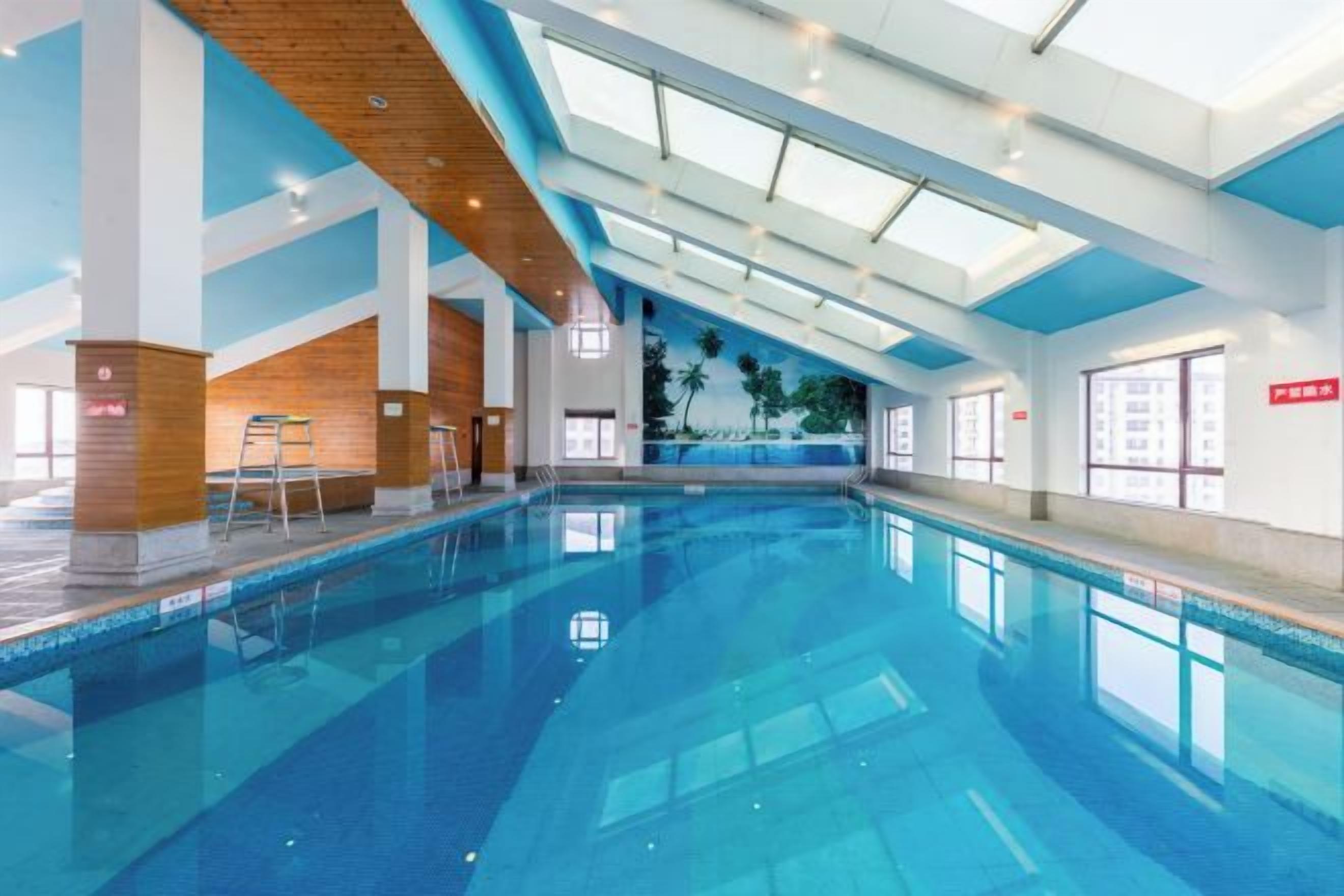An indoor swimming pool with clear blue water, wooden ceiling beams, and large windows allowing natural light.