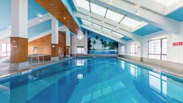 An indoor swimming pool with clear blue water, wooden ceiling beams, and large windows allowing natural light.