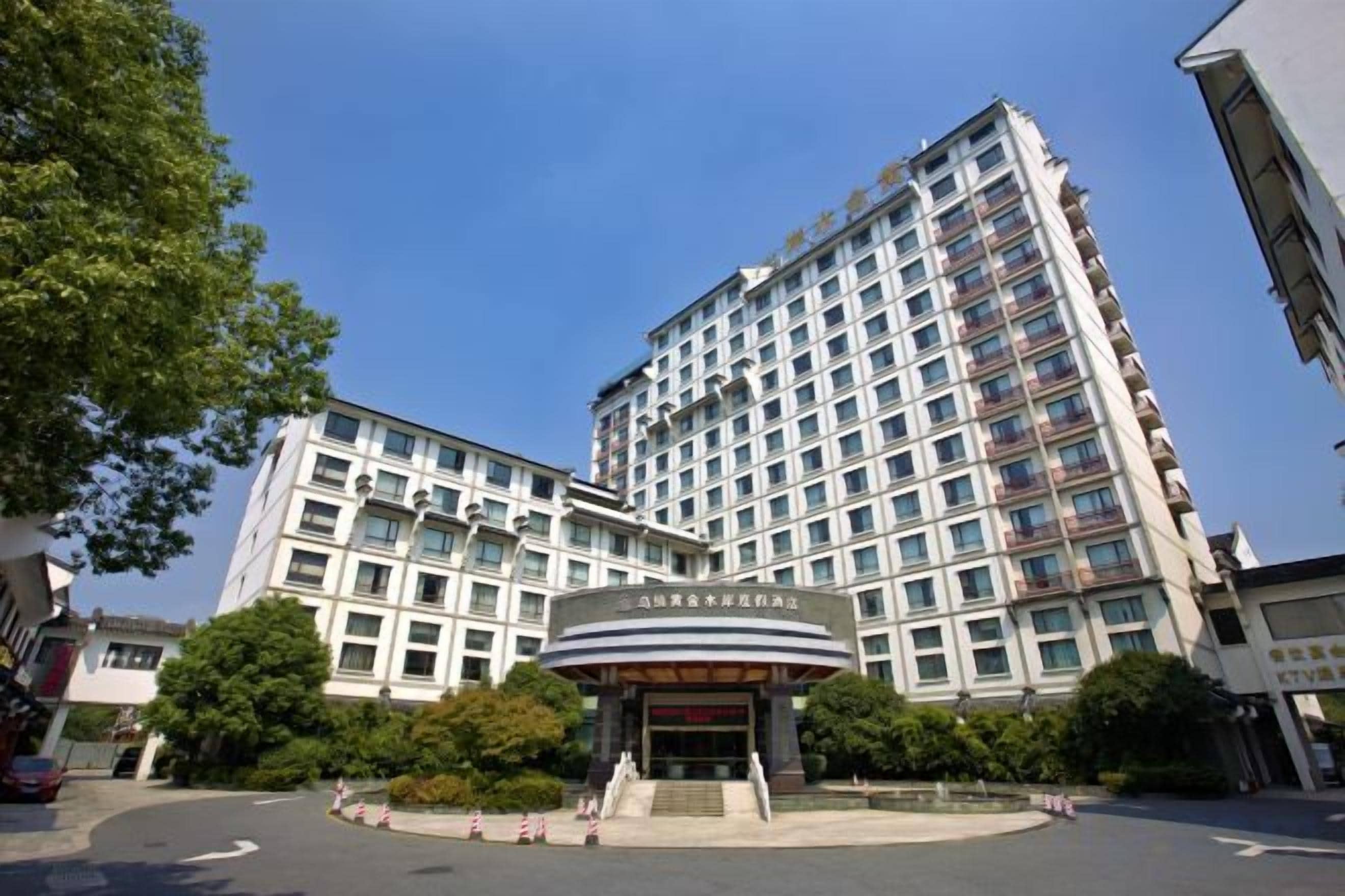 A multi-story hotel with a central entrance, surrounded by greenery and other buildings.