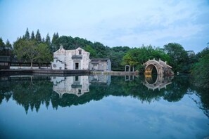 Um edifício tradicional refletido na água, uma ponte de pedra e árvores ao fundo.
