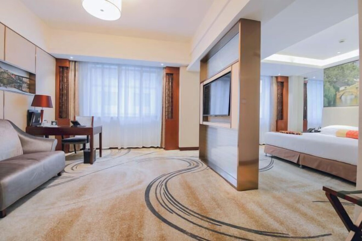 A hotel room with a sofa, a TV mounted on the wall, a bed, and a desk with a lamp.