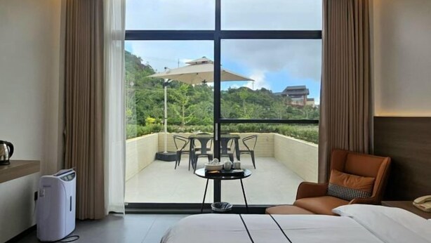 A hotel room with a bed, a table, a chair, and a view of a balcony with outdoor furniture. - Naixing Shihu Leisure Mountain Resort (Chaozhou)