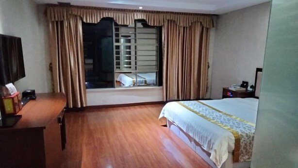 Una habitación de hotel con piso de madera, una cama con cubrecama blanco y amarillo, un televisor sobre un escritorio de madera y una ventana con cortinas.