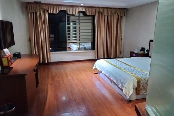 A hotel room with a wooden floor, a bed with a white and yellow bedspread, a television on a wooden desk, and a window with curtains. - Jiaoling Shun'an Hotel (Jiaoling)