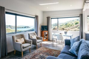 Un salon spacieux comprenant deux fauteuils, un canapé bleu, une table à manger avec des chaises et de grandes fenêtres donnant sur un paysage côtier.