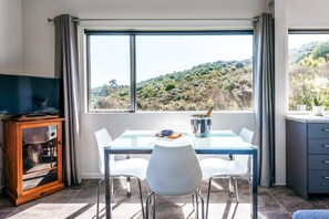 A dining area with a table, chairs, a television, and a view of a hilly landscape.