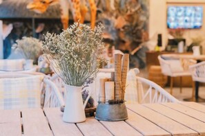Une table sur laquelle se trouve un vase blanc contenant une plante, un récipient en métal avec des papiers enroulés, et une table en bois, le tout dans un café.