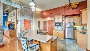 A kitchen with a brick wall, wooden cabinets, a stainless steel refrigerator, and a dining area with a table and chairs. - Downtown Cleveland Loft | Walk to Stadiums & Convention Center (Cleveland)