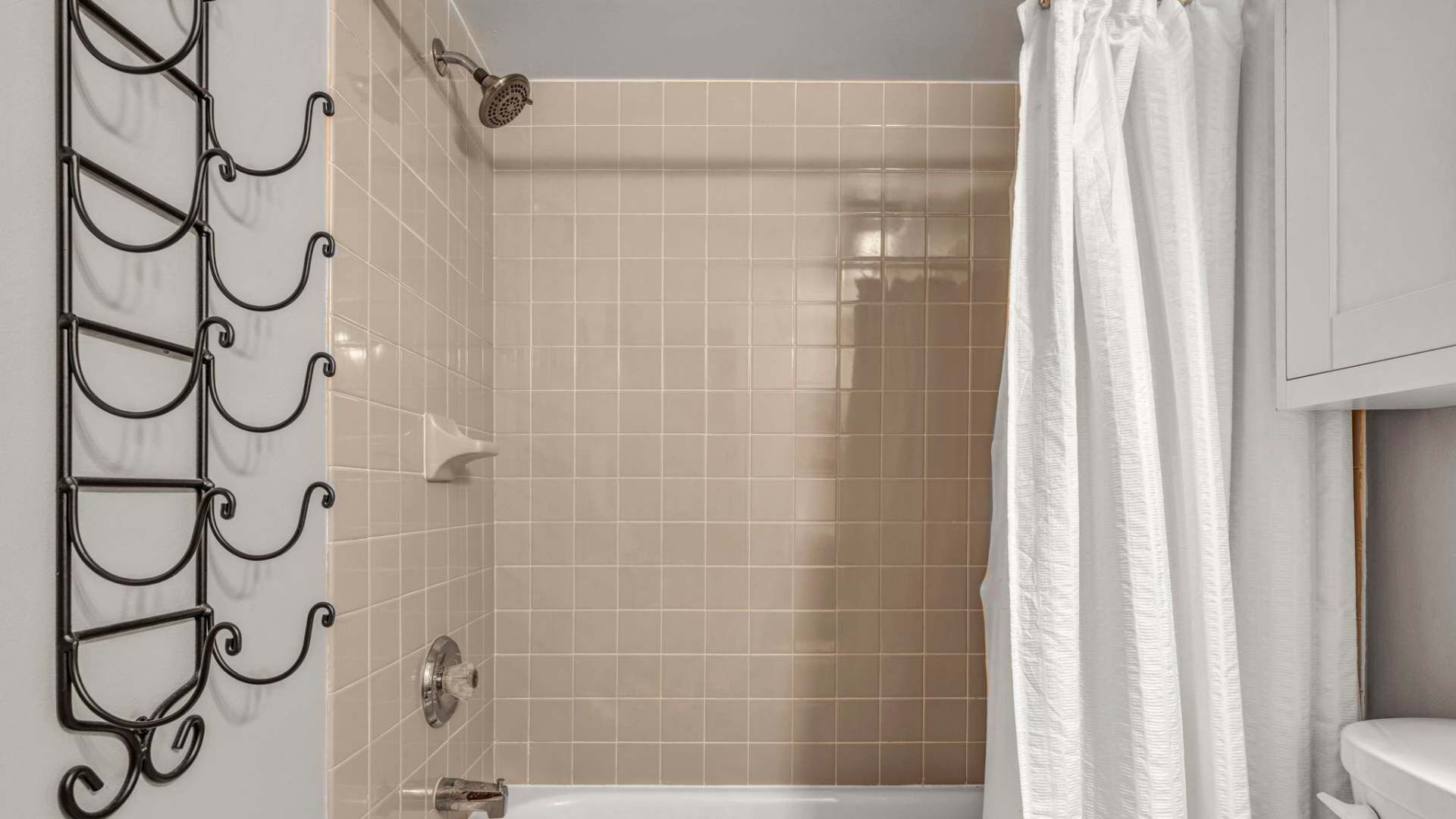 A bathroom with beige tiled walls, a white bathtub, a showerhead, and a metal rack with hooks.