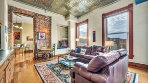 A spacious living room with a leather sofa, a coffee table, and a dining area with a brick wall and wooden flooring. - Downtown Cleveland Loft | Walk to Stadiums & Convention Center (Cleveland)