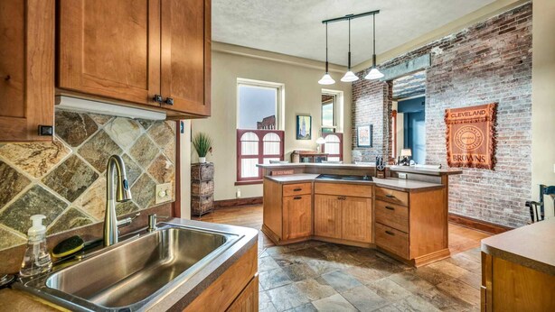 A kitchen with wooden cabinets, a stone backsplash, a brick wall, and pendant lighting. - Downtown Cleveland Loft | Walk to Stadiums & Convention Center (Cleveland)
