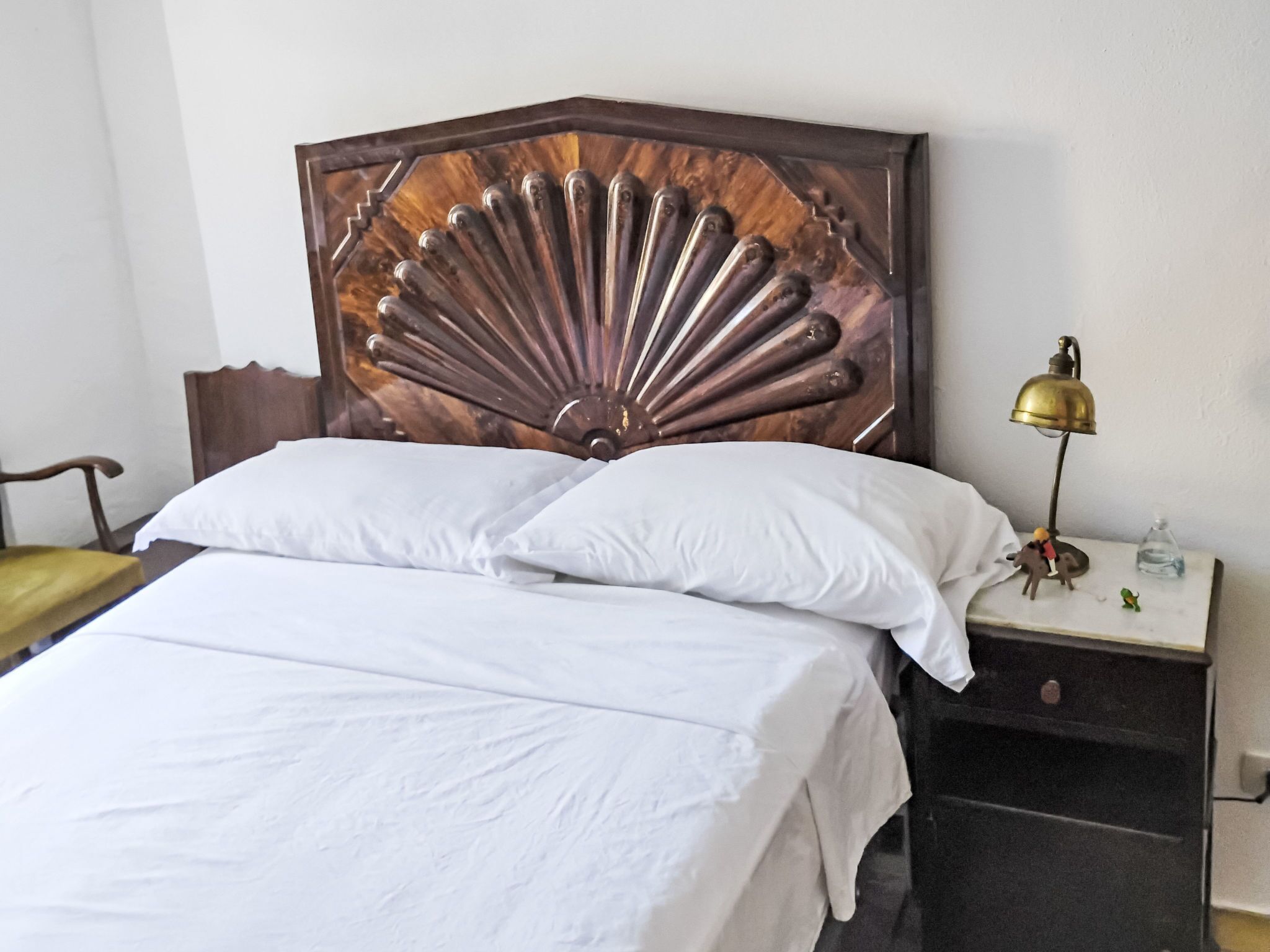 A neatly made bed with white linens, a wooden headboard with a fan-like design, a nightstand with a brass lamp, and a small figurine.