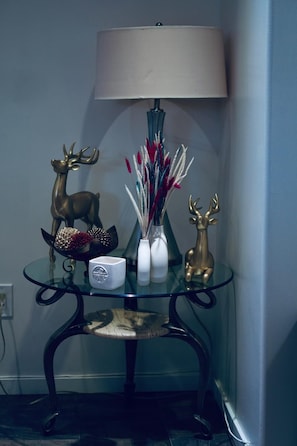 A serene corner setting with a glass-top table, featuring a decorative lamp, a vase with a floral arrangement, and two golden deer figurines.