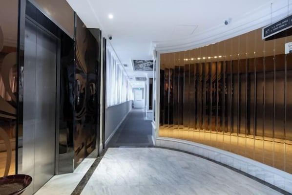 Un couloir doté d’un sol en marbre, d’un ascenseur en verre et d’un mur orné de stores verticaux.