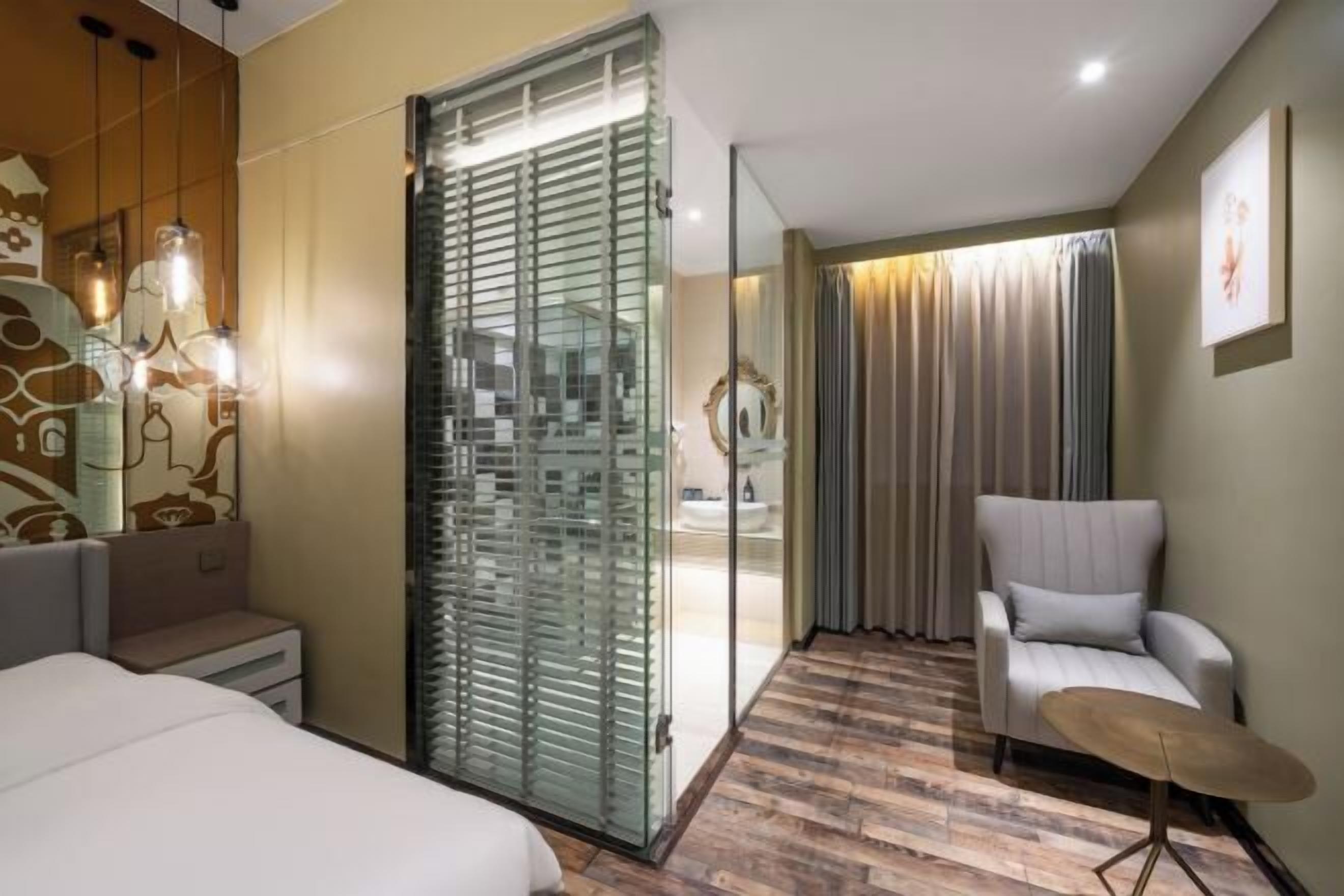 Quarto de hotel com uma cama, área de estar com uma cadeira, divisória de vidro e vista para um banheiro.