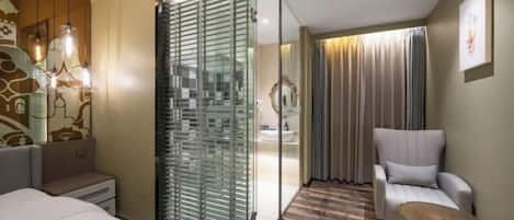 Habitación de hotel con cama, zona de estar con una silla, un panel de vidrio y vistas al baño.