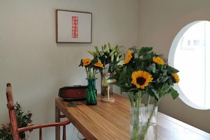 Una mesa de madera con jarrones de girasoles y una guitarra, un cuadro en la pared, y una ventana con vistas.