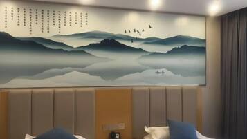Habitación de hotel con un gran mural en la pared que muestra montañas y pájaros, una cama con ropa blanca y almohadas azules, y una mesita de noche con un teléfono.