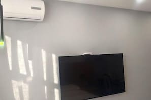 A room with a mounted flat-screen TV, a wall-mounted air conditioning unit, and a window casting shadows on the wall. - Yuanfang Hotel (Zhenglan Banner)