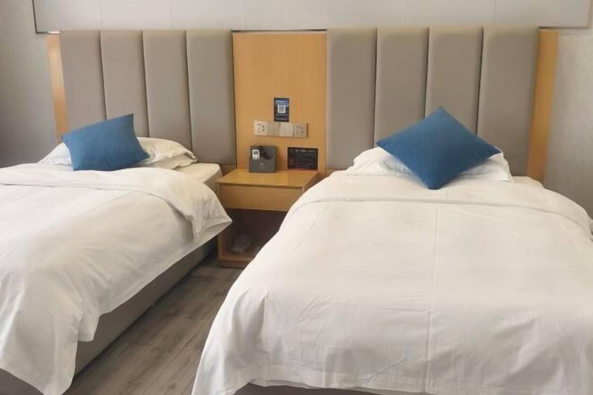 Een hotelkamer met twee eenpersoonsbedden, elk met een blauw kussen en wit beddengoed. Daartussen staat een houten nachtkastje met een kleine tafellamp en een stopcontact.
