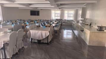 Un salón de conferencias con filas de mesas y sillas, una barra larga con electrodomésticos de cocina y una pantalla grande en la pared.