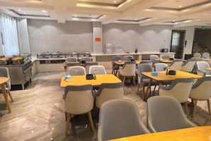 Un comedor de hotel con varias mesas y sillas, una cocina al fondo y una escalera que sube a un nivel superior.