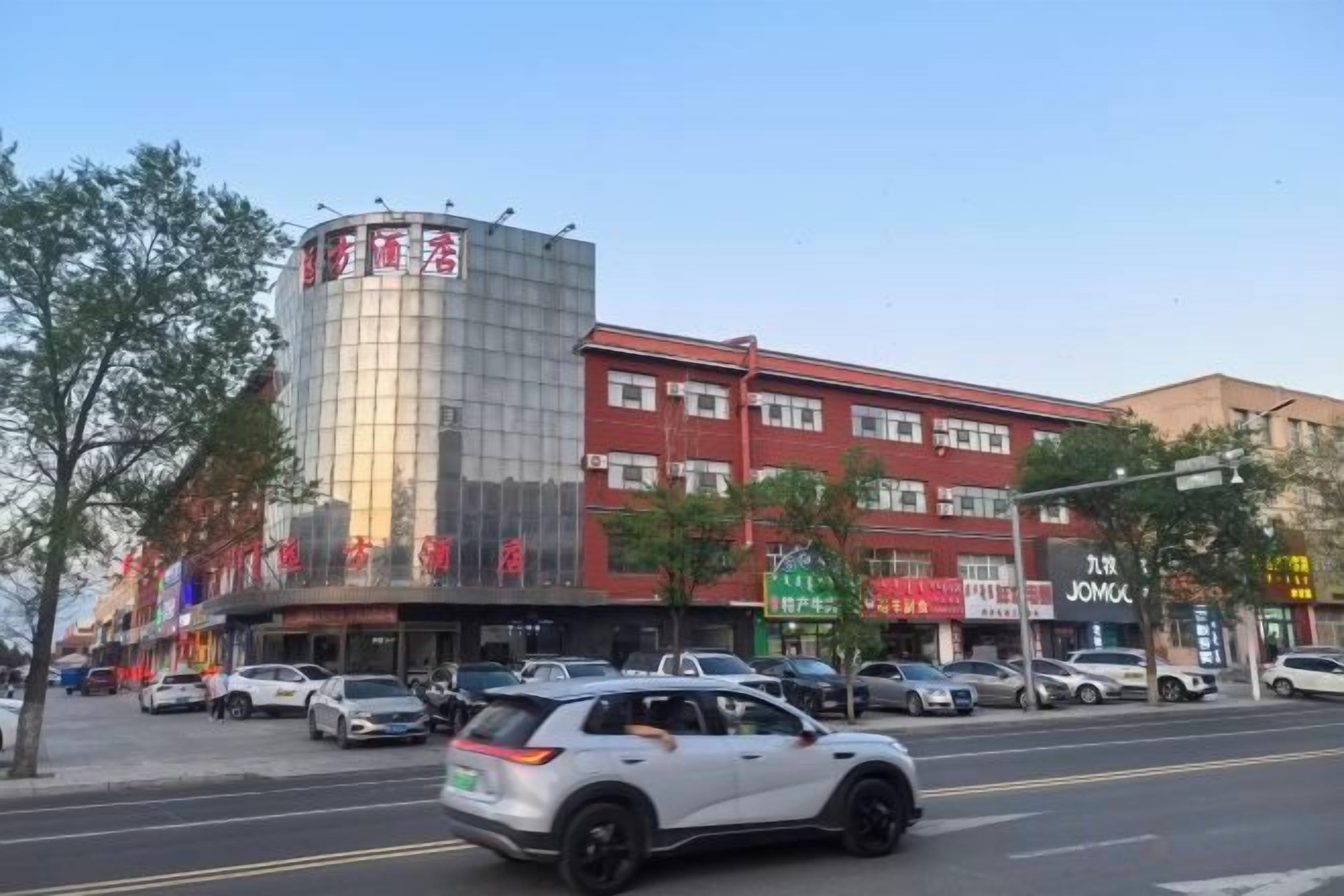 A multi-story building with commercial establishments on the ground floor, a parking lot in front, and a clear sky.