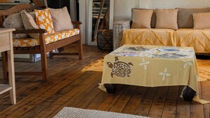 A cozy living room with a wooden floor, a sofa with floral cushions, a wooden chair, and a coffee table covered with a patterned tablecloth. - GREAT HOUSE BY MANAEVA LODGE (Moorea-Maiao)