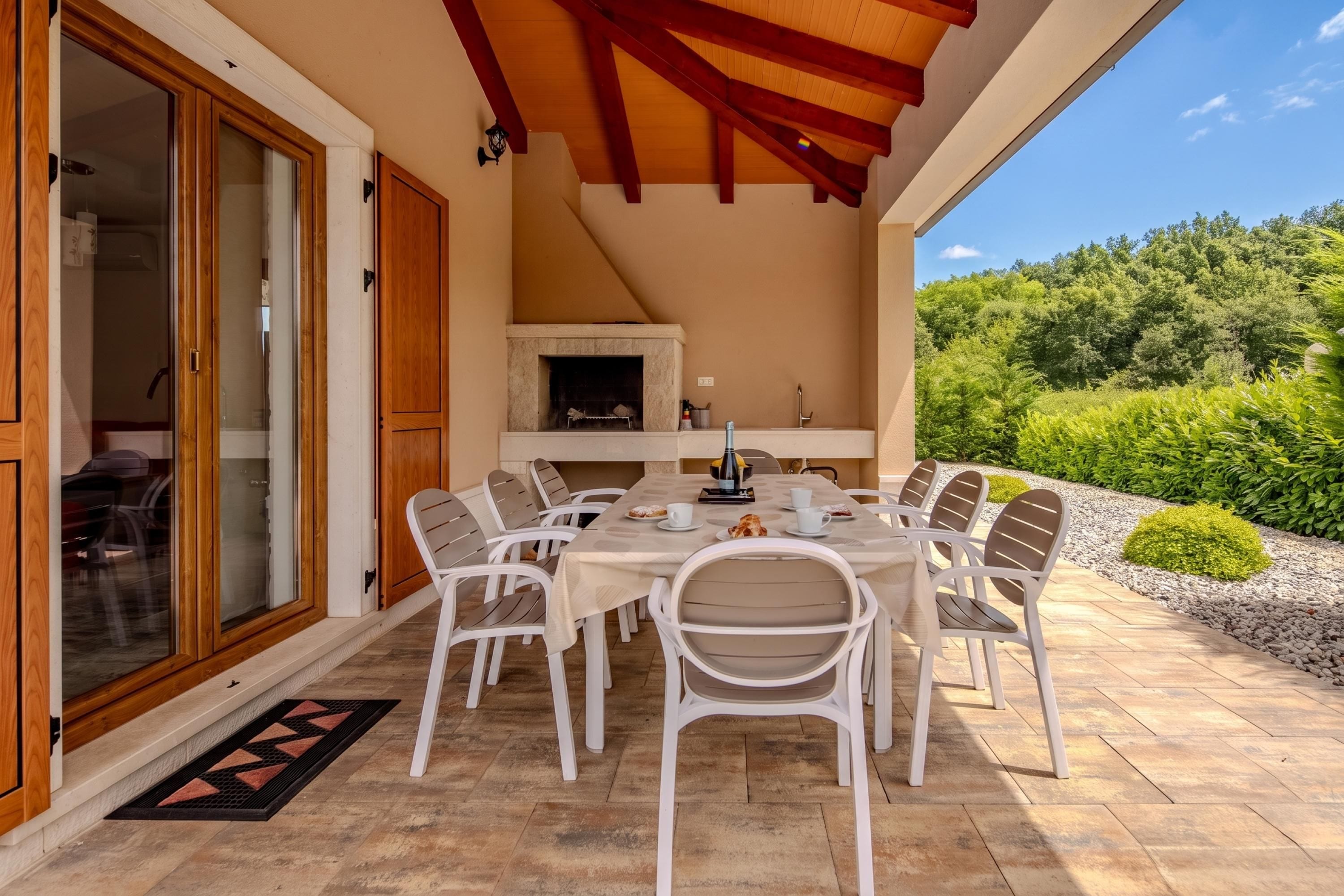 A covered patio with a dining table and chairs, a fireplace, and a view of greenery.