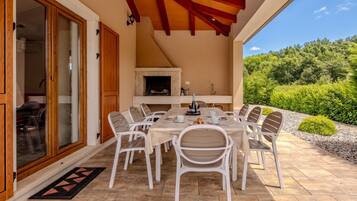 A covered patio with a dining table and chairs, a fireplace, and a view of greenery.