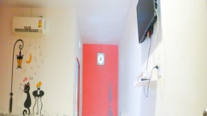 A hotel room with a bed, a wall-mounted TV, an air conditioner, a red accent wall, and a tiled floor.