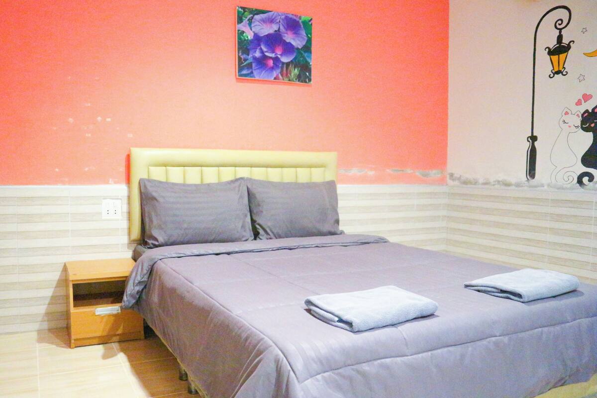 A bedroom with a bed, pillows, a nightstand, a wall-mounted lamp, and a small picture of flowers.