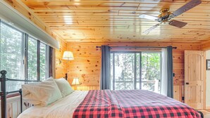 Quarto de cabana de madeira com cama, ventilador de teto e janelas amplas.