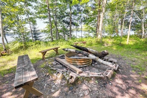 A rustic campsite with a wooden table, benches, and a fire pit surrounded by trees and a lake.
