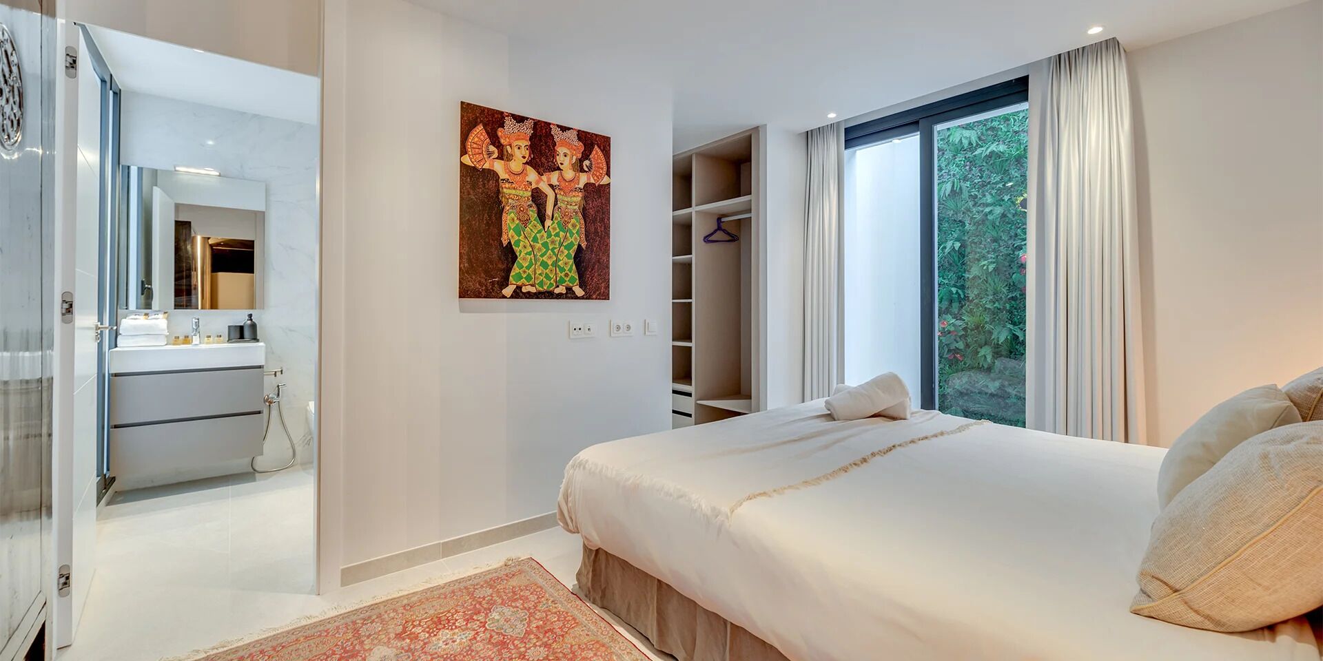 A modern bedroom with a large bed, a walk-in closet, a bathroom with a mirror, and a view of greenery through the window.