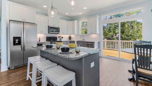 A modern kitchen with white cabinets, a stainless steel refrigerator, and a large island with stools. - Great Location Near Beach, Walk to Restaurants and shops, Fenced in Back Yard (Oak Island)