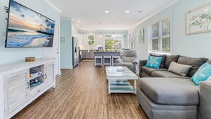 A spacious living area with a grey sofa, a coffee table, and a TV mounted on the wall. - Great Location Near Beach, Walk to Restaurants and shops, Fenced in Back Yard (Oak Island)