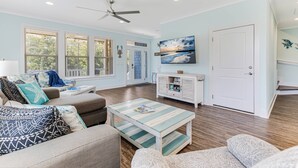 A spacious living room with a ceiling fan, large windows, a coffee table, and a door leading to another room. - Great Location Near Beach, Walk to Restaurants and shops, Fenced in Back Yard (Oak Island)