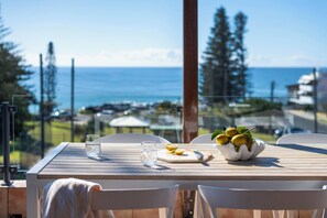 A wooden table with a bowl of lemons and a glass of water, overlooking a scenic view of the ocean and trees. - Indigo Terrace One - Oceanstays- Pet Friendly (Yamba)