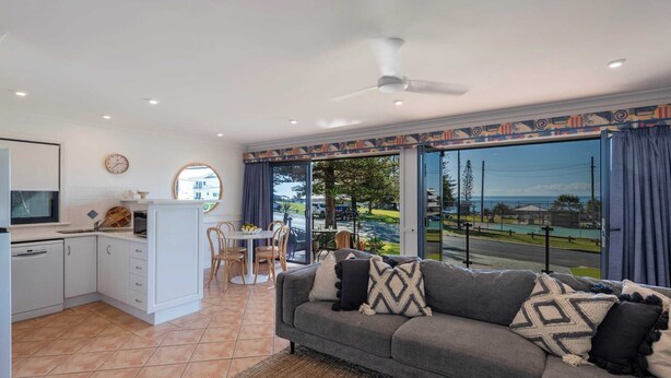 A modern living room with a grey sofa, a dining area with a table and chairs, a kitchen with white cabinets, and large windows offering a view of the outdoors.