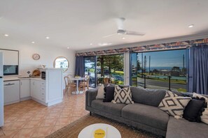 A modern living room with a grey sofa, a dining area with a table and chairs, a kitchen with white cabinets, and large windows offering a view of the outdoors.
