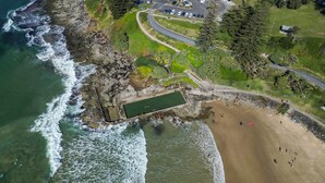 An aerial view of a coastal area with a rocky shoreline, a swimming pool, and a sandy beach.