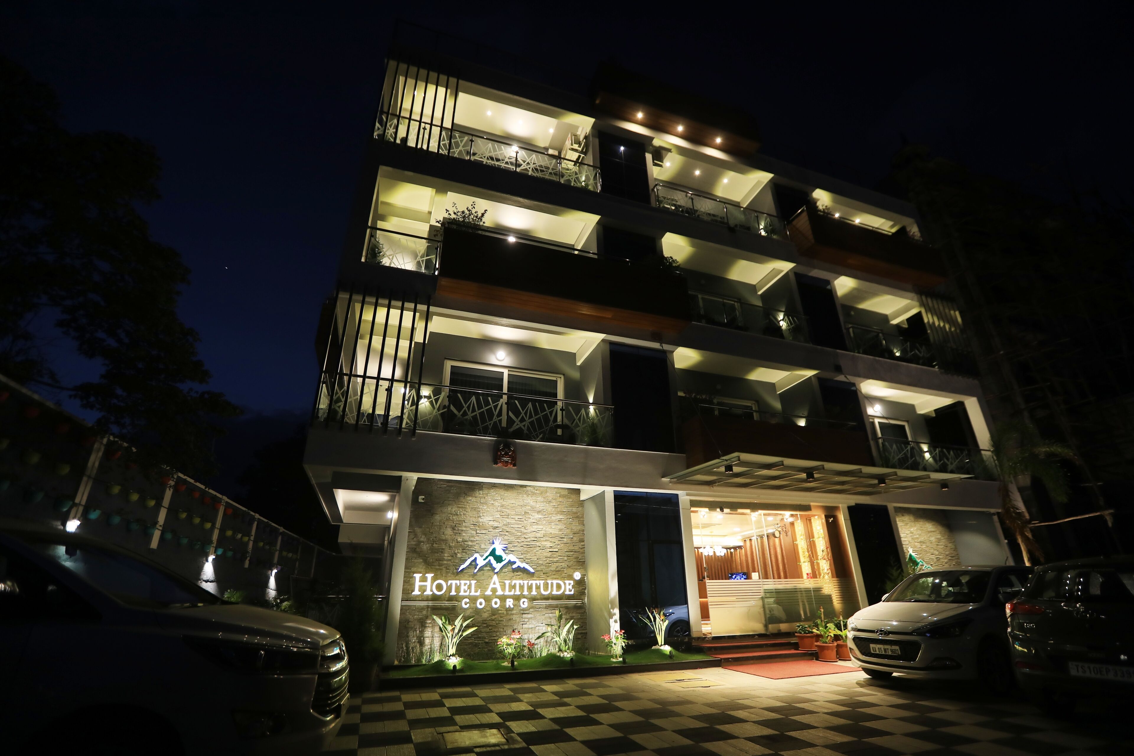 Un hôtel à plusieurs étages, doté d’une entrée à carreaux, de balcons et d’une enseigne indiquant « Hôtel Altitude Coorg ».