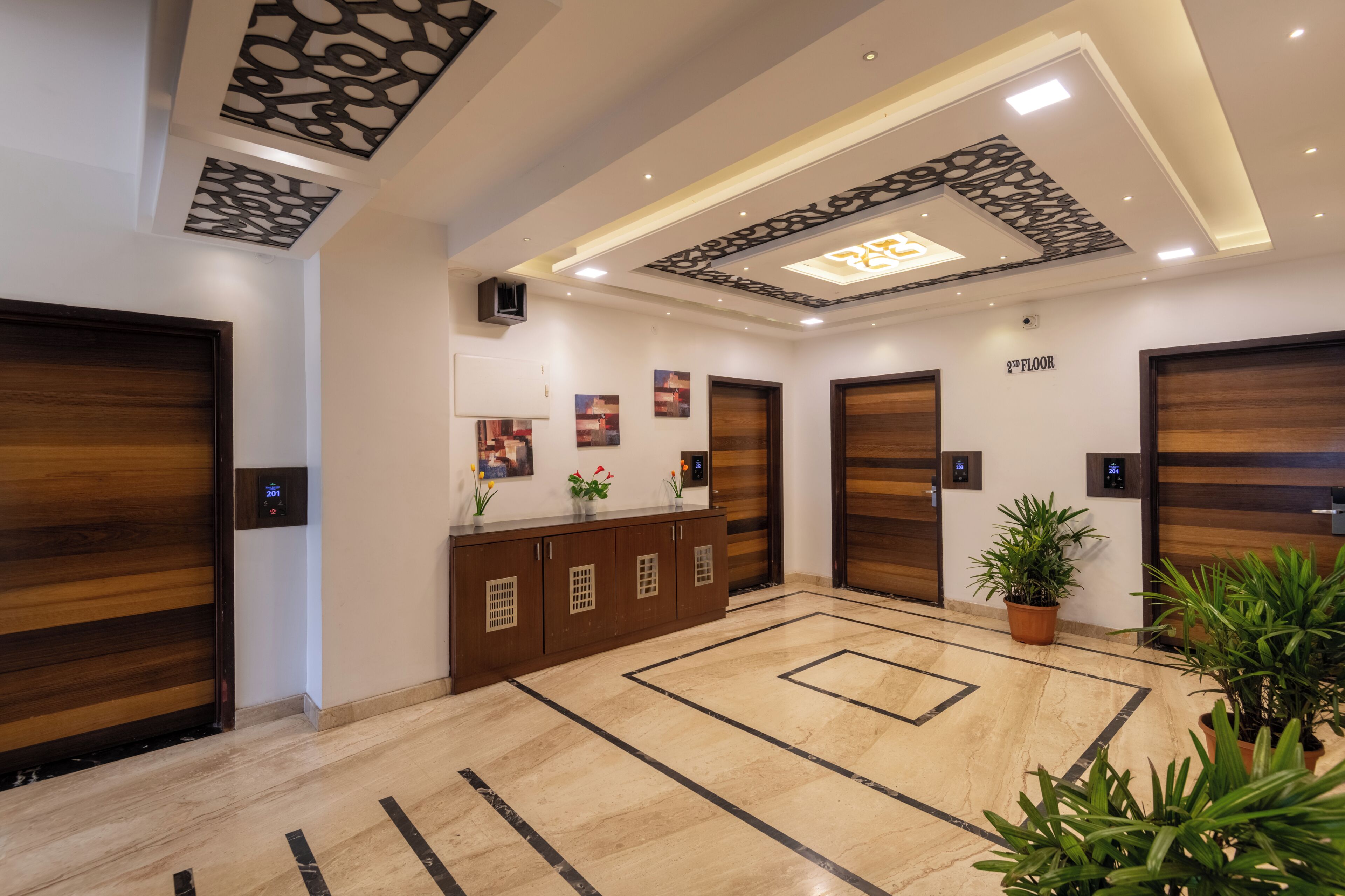 A modern lobby with a reception desk, two wooden doors, a ceiling with decorative panels, and a potted plant.
