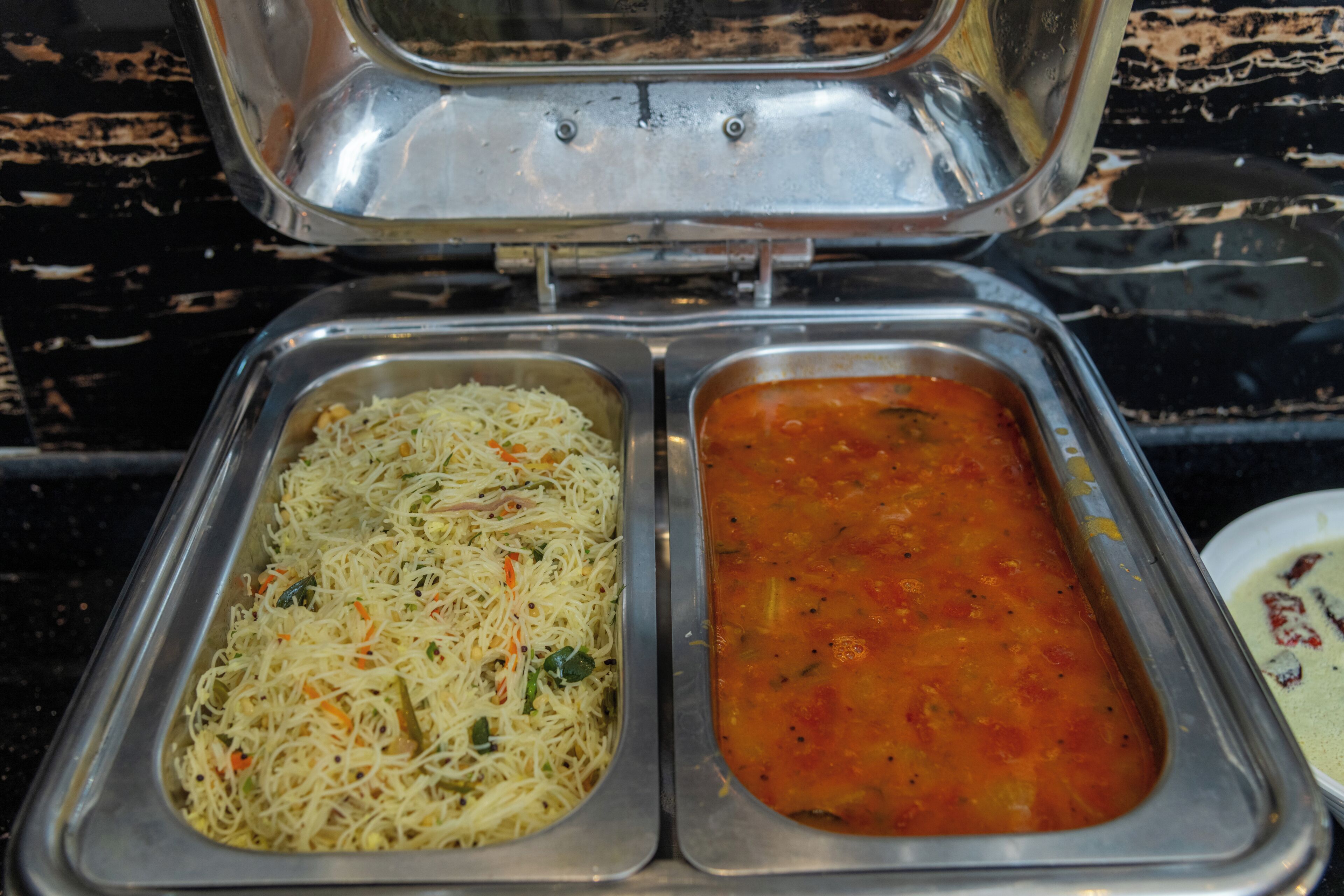 A stainless steel buffet with two compartments, one containing noodles and the other a tomato-based soup.